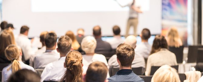 The crowd at a conference is sat listening to a speaker in the background.