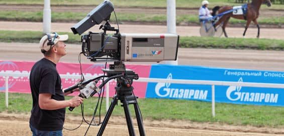 A camera man with a camera designed for outdoor broadcasting at a horse racing related event.