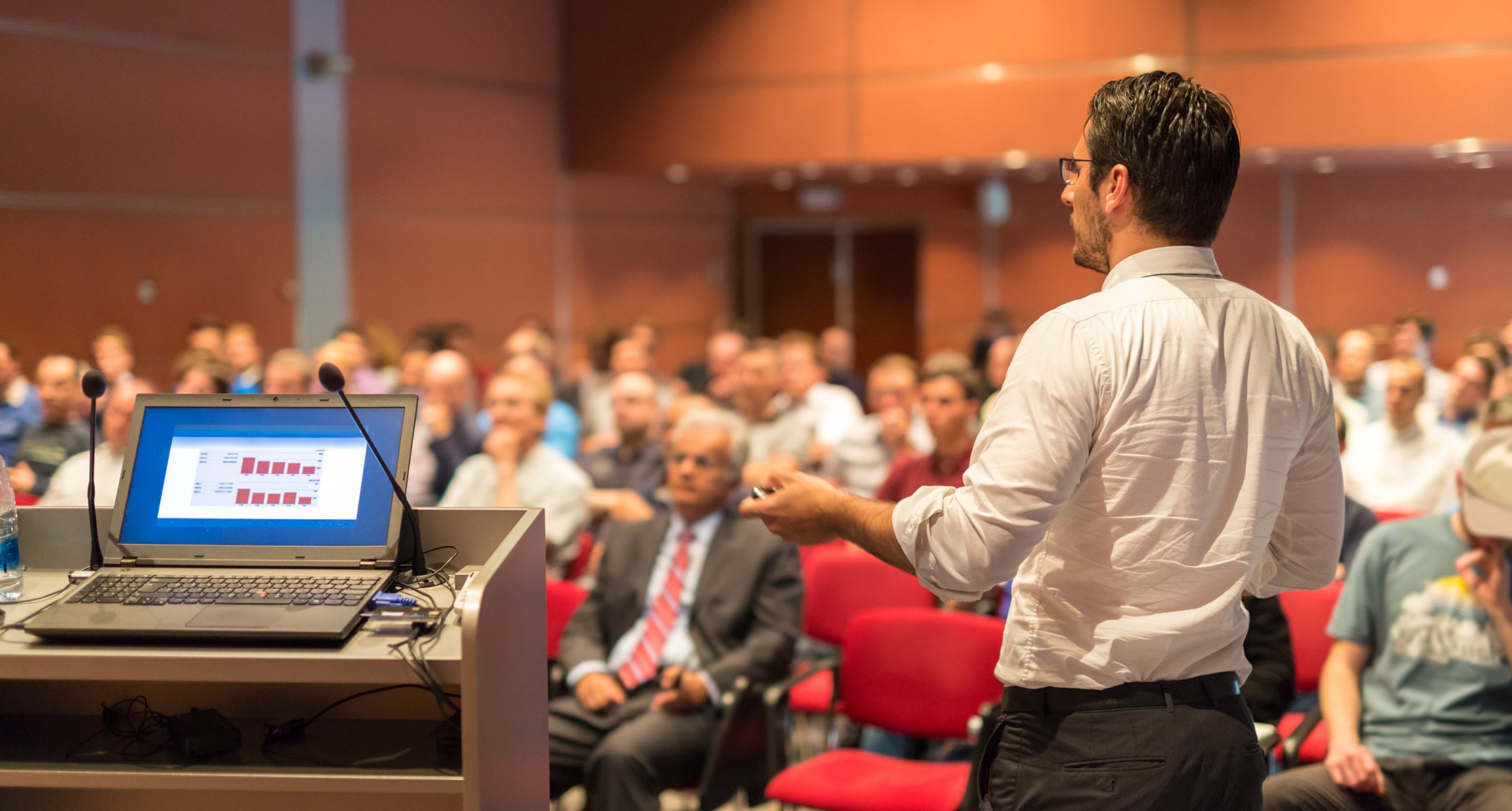 A speaker at a conference in the foreground his seated audience in the background.
