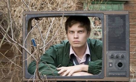 A slightly grumpy looking man looking through the shell of a broken television without a screen.