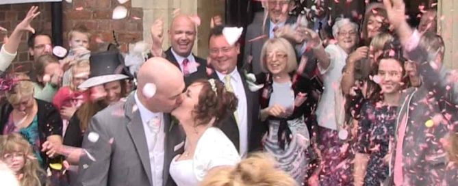 A newly wedded couple leaving the church. Confetti is being thrown by the crowd that surrounds them.
